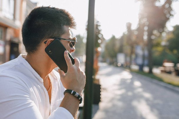 νεαρός κομψός τύπος σε πουκάμισο με τηλέφωνο στον πάγκο για ηλιόλουστη ζεστή μέρα σε εξωτερικούς χώρους - Φωτογραφία, εικόνα