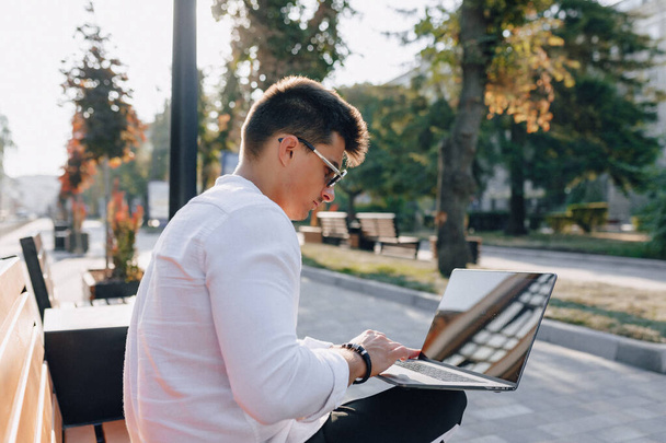 young stylish guy in shirt with phone and notebook on bench on sunny warm day outdoors, freelance - Photo, Image