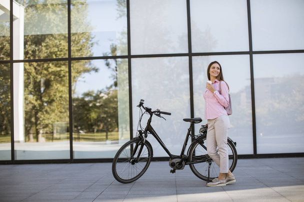 Mooie jonge vrouwelijke fietser drinkt koffie uit een kopje bij de elektrische fiets in de stedelijke omgeving - Foto, afbeelding