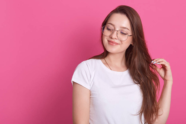 Portrait of happy young cute brown haired girl sytands smiling and looking aside, touches hair, posing isolated on pink background, looks thouthfull, dreams about something. Copyspace for advertisment - Foto, Bild