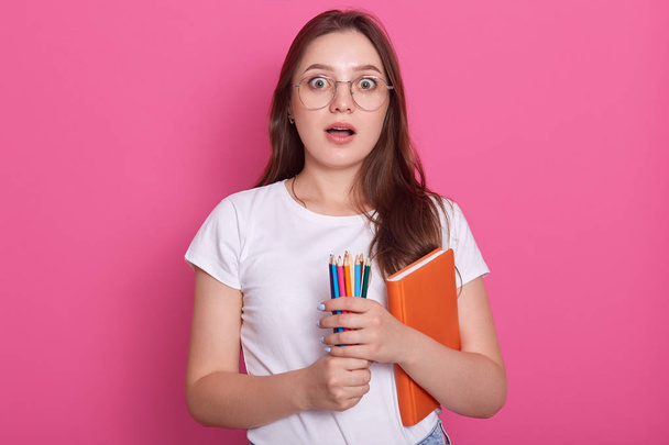 Astonished dark haired woman with opened mouth, carries textbook and colored pencils for writing or drawing, being ready to make notes while listens some shocking information, poses on pink background - Foto, afbeelding