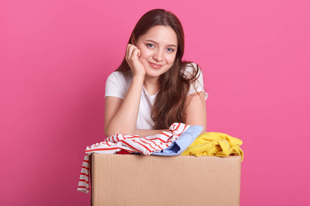 Donation Concept. Woman holding donate box with full of clothes, adorable smiling female keeps hand under chin and clothes donate box, lady wearing casual white t shirt, posing isolated on pink. - Photo, image