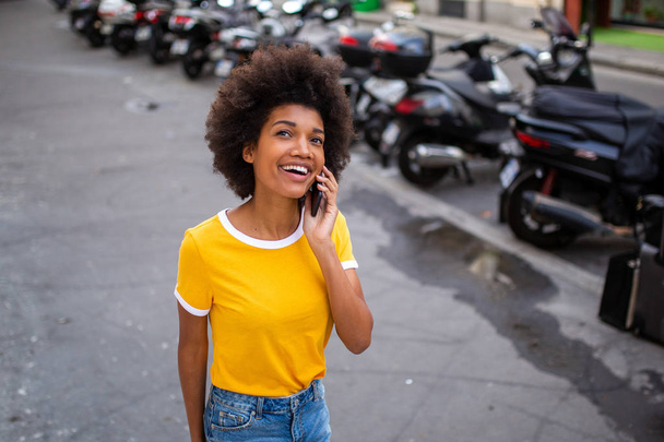 Портрет счастливой африканской молодой женщины, идущей и разговаривающей с мобильным телефоном на улице
 - Фото, изображение