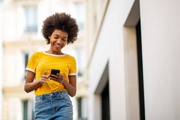 Portrait de belle jeune femme afro-américaine heureuse marchant avec un téléphone portable en ville
 - Photo, image