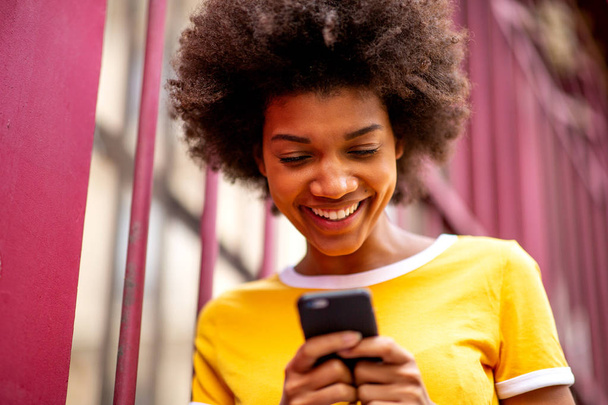 Zbliżenie portret uśmiechniętej, młodej, afrykańskiej kobiety patrzącej na telefon komórkowy na zewnątrz - Zdjęcie, obraz