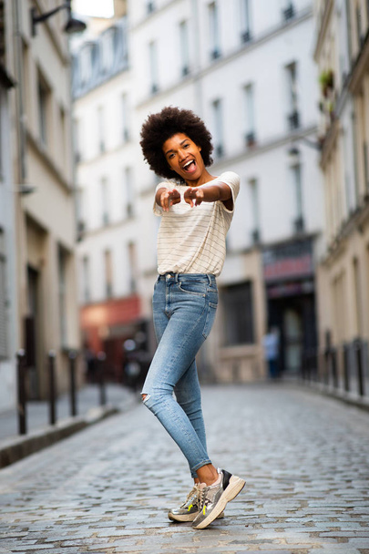 Full body portrait of cheerful young african american woman standing on city street with pointing - Fotó, kép
