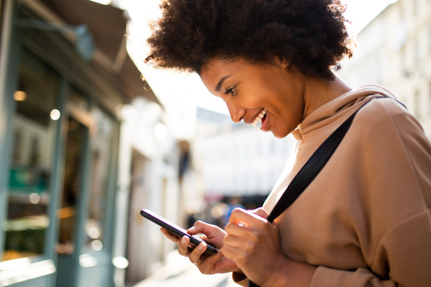 Side portrait of happy young black woman looking at mobile phone in city - Photo, Image