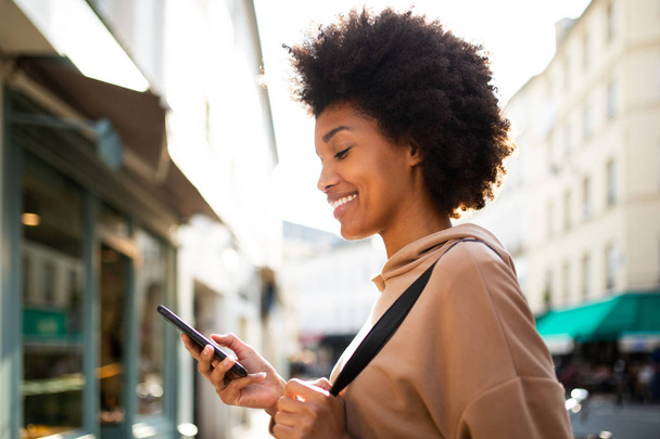 Boczny portret uśmiechniętej czarnej kobiety patrzącej na telefon komórkowy w mieście - Zdjęcie, obraz