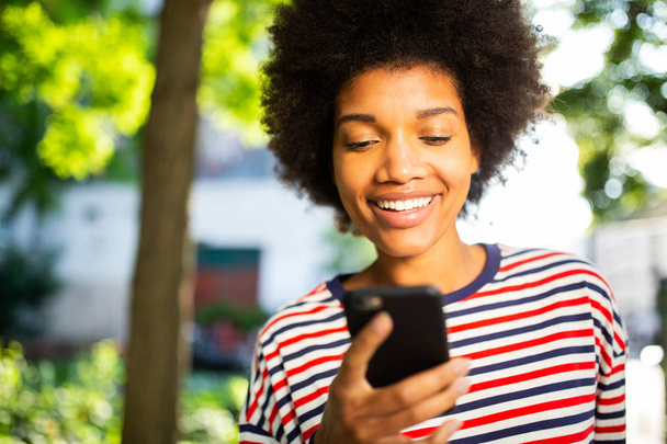 Close up portrait beautiful young black woman with afro hair smiling at mobile phone in park  - 写真・画像