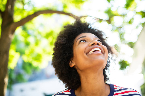 Крупним планом портрет красива молода чорна жінка посміхається з афро волоссям в парку і дивиться вгору
 - Фото, зображення