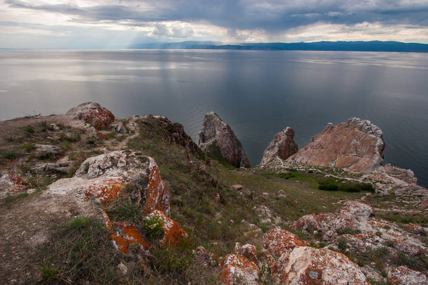 Sziklák Három Testvér Olkhon szigetén a Bajkál-tónál. Égbolt felhőkkel. A köveken vörös moha és zöld fű van. Esik a távolban, hegyek láthatók.. - Fotó, kép