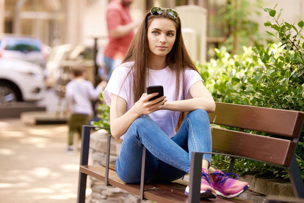 Νεαρή γυναίκα μήνυμα κειμένου, ενώ κάθεται στον πάγκο στην str - Φωτογραφία, εικόνα
