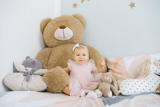 una niña de un año en un vestido rosa se sienta en la cama rodeada de juguetes suaves y almohadas, ella es feliz y sonriente
 - Foto, Imagen