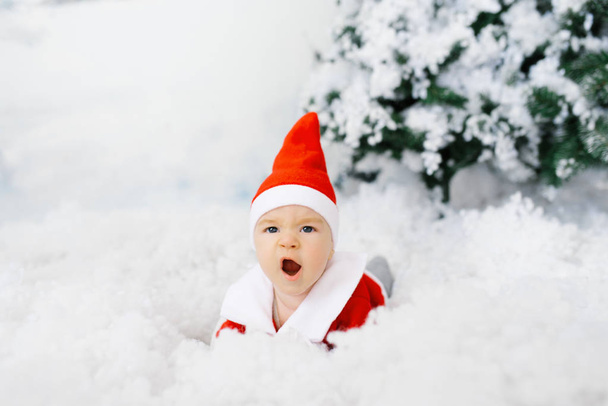 The kid in the clothes of little Santa is lying on his stomach on the artificial snow, he is surprised. Christmas sales concept for kids - Photo, Image
