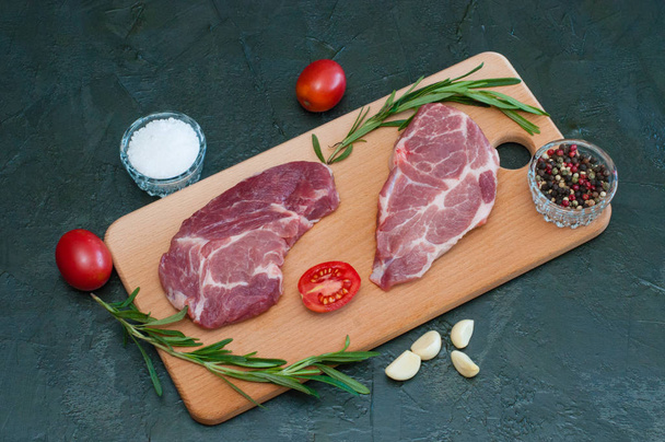 Fladen, Schweinefleisch, rohe Steaks und Gewürze, Knoblauch, Salz, Pfeffer, geschnittene Tomaten und Rosmarin auf Schneidebrett - Foto, Bild