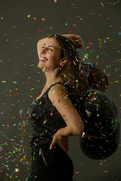 Attractive woman holding black baloons and dancing under falling confetti, party and holiday season concept - Photo, Image