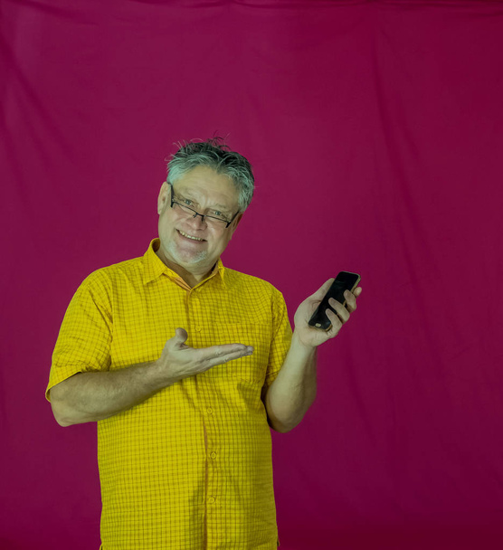 Een oudere man met grijs haar houdt een smartphone in zijn handen. Verschillende emoties worden op het gezicht geuit. Vreugde, plezier, obscuriteit, bedachtzaamheid, genot, tederheid. Portret van een man in een geel shirt op een rode achtergrond met een telefoon. - Foto, afbeelding