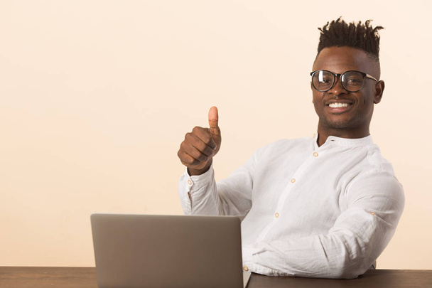 handsome young african man in glasses and a white shirt sits at a table with a laptop on a beige background - Фото, изображение