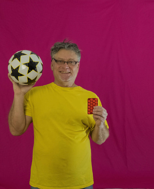 Un anciano con el pelo gris sostiene un voleibol en sus manos. Diferentes emociones se expresan en la cara. Retrato de un hombre con una camiseta amarilla sobre un fondo rojo. Deportes contra las drogas
. - Foto, imagen