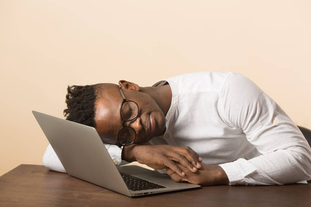 handsome young african man in glasses and a white shirt sits at a table with a laptop on a beige background - Foto, afbeelding
