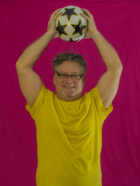 Un anciano con el pelo gris sostiene un voleibol en sus manos. Diferentes emociones se expresan en la cara. Retrato de un hombre con una camiseta amarilla sobre un fondo rojo. Deportes contra las drogas
. - Foto, imagen