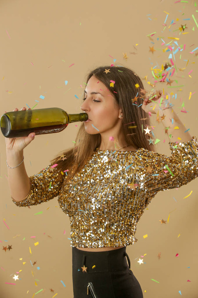 Atrakcyjna kobieta w złotej koszuli z cekinami tańcząca i pijąca wino z butelki. Szczęśliwa młoda i piękna kobieta korzystająca z imprezy z confetti. - Zdjęcie, obraz