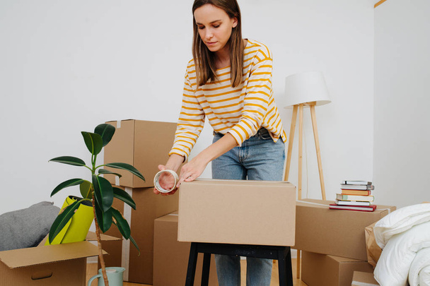 Woman sealing cardboard box, she's moving out from old apartment to a new one. - Foto, Bild