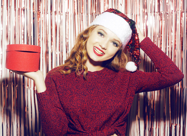 Menina bonita em um chapéu de Papai Noel brilhante em um fundo brilhante detém
 - Foto, Imagem
