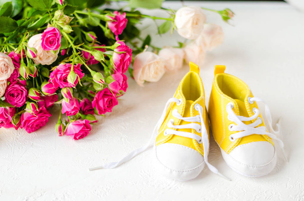 Żółte buty dziecięce, stojące na białym stole z różami. zbliżenie. koncepcja odzieży dziecięcej. Pozdrowienia dla dzieci w żółtych butach  - Zdjęcie, obraz