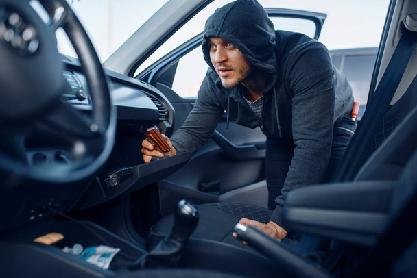 Car robber takes the wallet from the glove compartment, criminal lifestyle, stealing. Hooded male bandit opening vehicle on parking. Auto robbery, automobile crime - Foto, Imagem