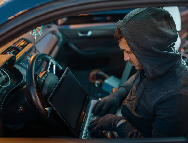 Profesjonalny złodziej samochodów z laptopem hakujący system bezpieczeństwa, przestępczy styl życia. Zakapturzony złodziej otwierający samochód na parkingu. Rabunek samochodowy, przestępstwa samochodowe - Zdjęcie, obraz