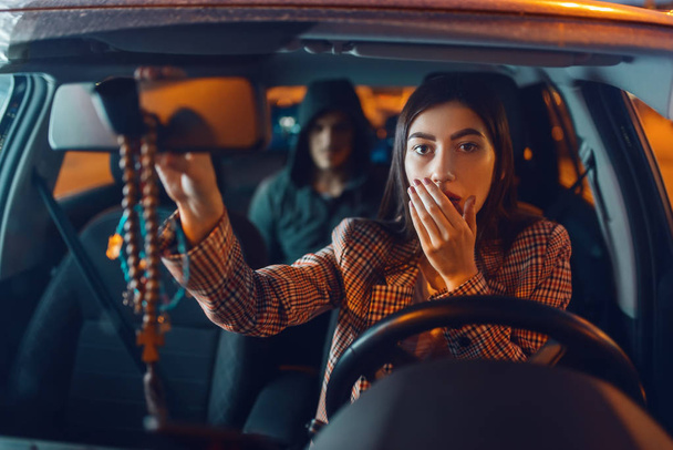 Female driver and hooded car hijacker on back seat, criminal lifestyle, stealing. Male bandit and victim in vehicle on parking. Auto robbery, automobile crime, hijacking - 写真・画像