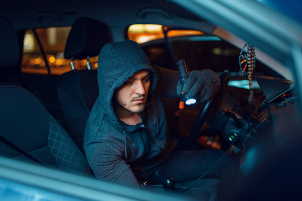 Profesionální zloděj aut s baterkou, kriminálním životním stylem, krádeží. Muž s kápí hledající zboží ve vozidle na parkovišti, kradl. Loupež v autě, automobilová kriminalita - Fotografie, Obrázek