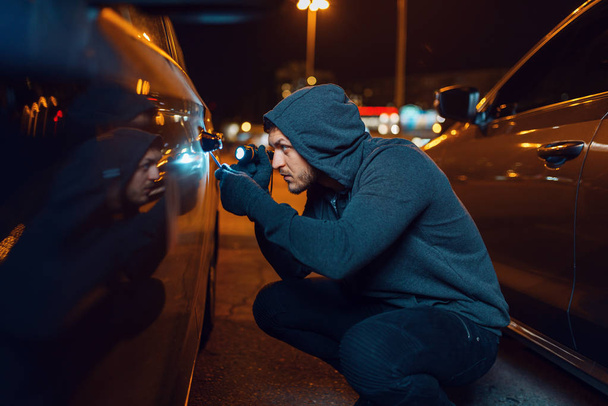 Автоугонщик со взломанной отверткой. Мужчина в капюшоне открывает машину на парковке. Ограбление автомобилей, автомобильные преступления
 - Фото, изображение