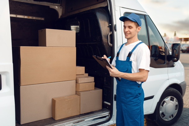 Deliveryman in uniform, carton boxes in the car, delivery service. Man standing at cardboard packages in vehicle, male deliver, courier or shipping job - Фото, изображение