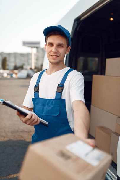 Deliveryman in uniform gives parcel, carton boxes in the car, delivery service. Man standing at cardboard packages in vehicle, male deliver, courier or shipping job - 写真・画像