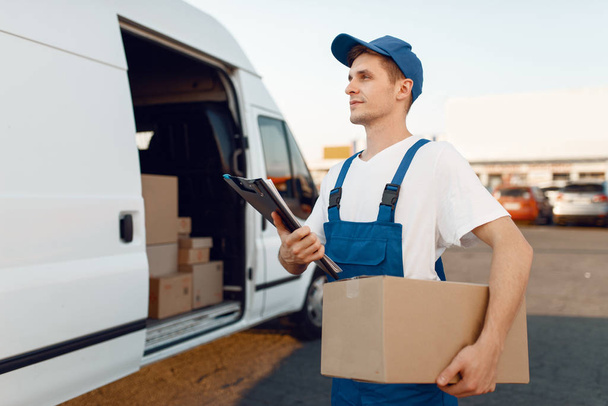 Deliveryman in uniform gives parcel, carton boxes in the car, delivery service. Man standing at cardboard packages in vehicle, male deliver, courier or shipping job - Фото, изображение