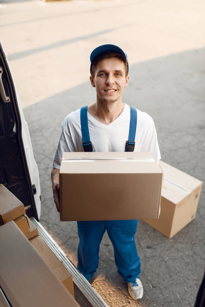 Deliveryman in uniform unloads the car with parcels, delivery service. Man standing at cardboard packages in vehicle, male deliver, courier or shipping job - Foto, Bild