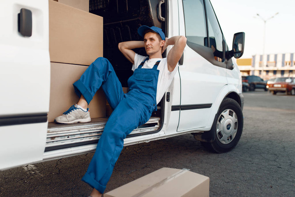 Deliveryman in uniform relaxing in the car during a break, auto with parcels and carton boxes, delivery service. Man poses at cardboard packages in vehicle, male deliver, courier or shipping job - Photo, Image
