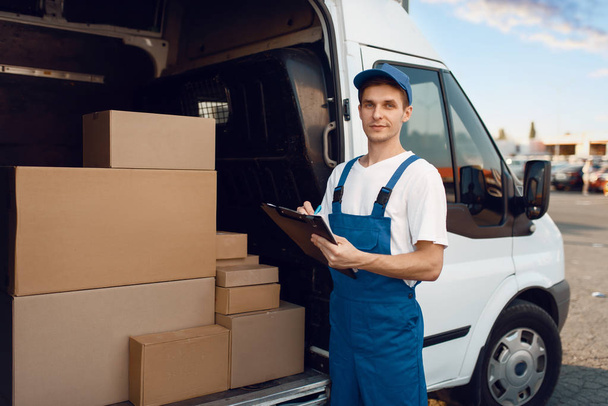 Deliveryman in uniform, carton boxes in the car, delivery service. Man standing at cardboard packages in vehicle, male deliver, courier or shipping job - Photo, Image