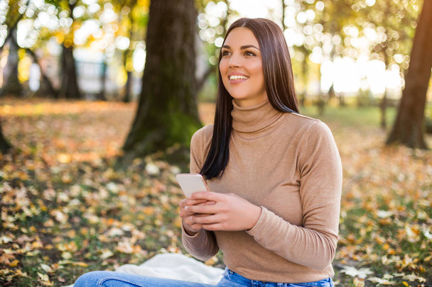 Beautiful woman texting messages while sitting in the park and enjoys in autumn. - 写真・画像