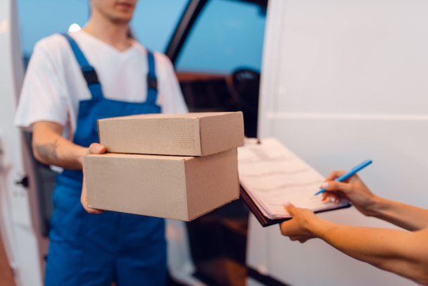 Deliveryman in uniform gives parcel to female recipient at the car, delivery service. Man holding cardboard package near the vehicle, male deliver and woman, courier or shipping job - Photo, Image