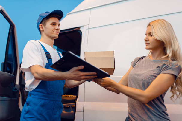 Deliveryman in uniform gives parcel to female customer at the car, delivery service. Man holding cardboard package near the vehicle, male deliver and woman, courier or shipping job - Valokuva, kuva