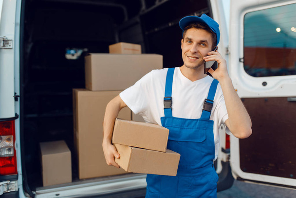 Deliveryman in uniform holds parcel and mobile phone at the car, delivery service, delivering. Man standing at cardboard packages in vehicle, male deliver, courier or shipping job - Photo, image