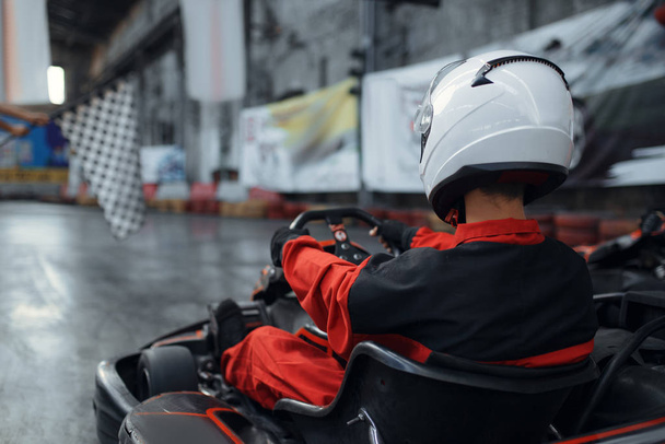 Kart racer in helmet, back view, karting auto sport indoor. Speed race on close go-cart track with tire barrier. Fast vehicle competition, high adrenaline leisure - Foto, afbeelding