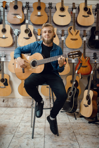 Мужской гитарист играет на акустической гитаре в музыкальном магазине. Ассортимент в магазине музыкальных инструментов, оборудование для покупки музыкантов
 - Фото, изображение