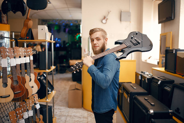 Young man poses with electric guitar on his shoulder in music store. Assortment in musical instruments shop, male musician buying equipment - Photo, Image