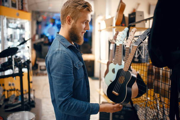 Bearded young man choosing ukulele guitar in music store. Assortment in musical instruments shop, male musician buying equipment - Photo, Image