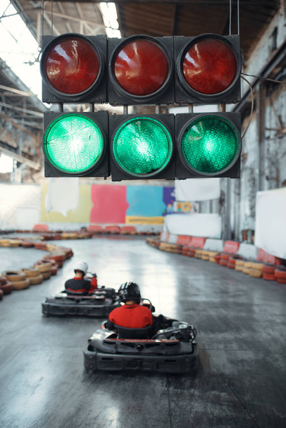 Two kart racers starts race, green traffic light, back view, karting auto sport indoor. Speed race on close go-cart track with tire barrier. Fast vehicle competition, high adrenaline leisure - Photo, Image