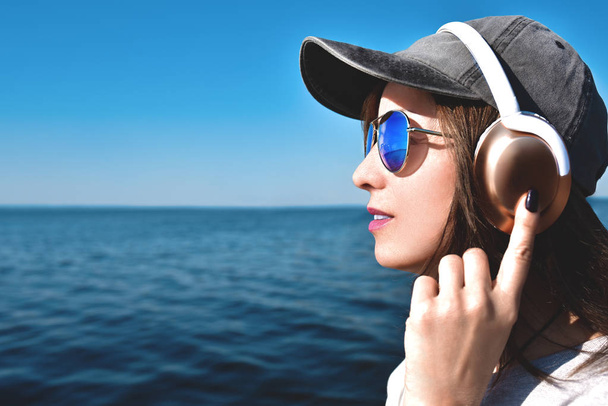 close portrait of a woman in sunglasses wearing a cap who likes music on the background of the sea.Muziekconcept. - Foto, afbeelding